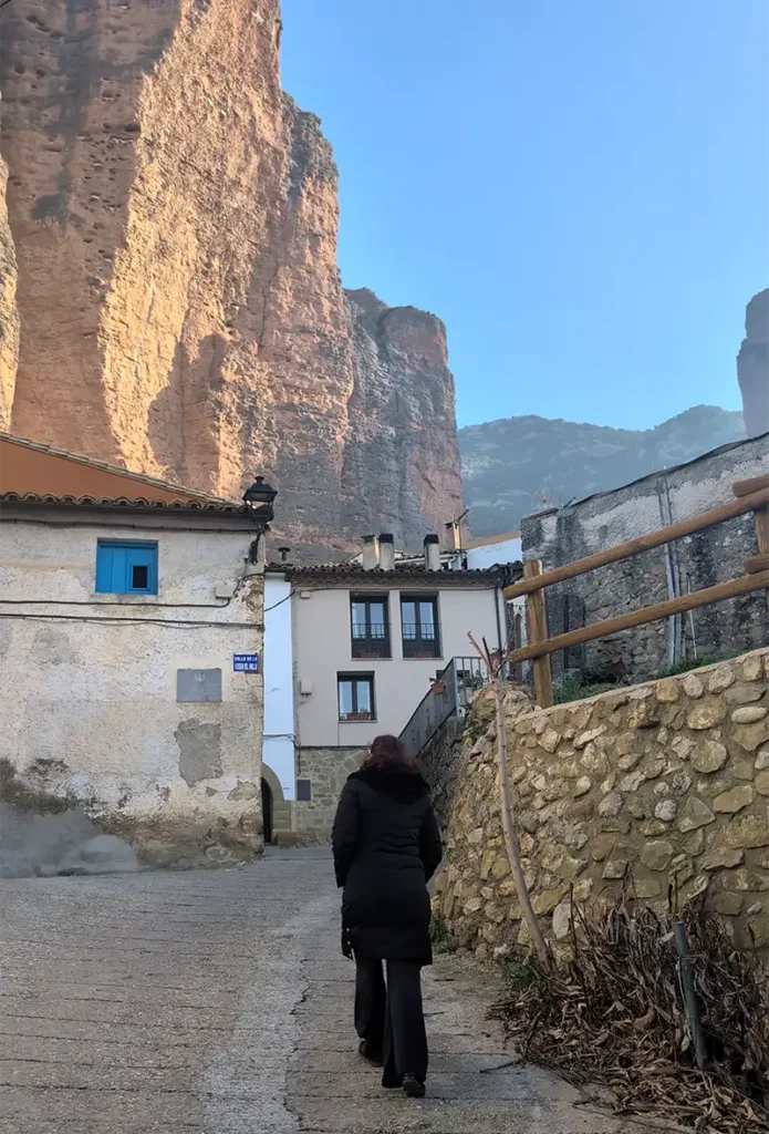 Mulher caminhando por uma ruela de Riglos com os mallos, paredões de rocha vermelha, dominando o horizonte ao fundo, Huesca, Aragão.