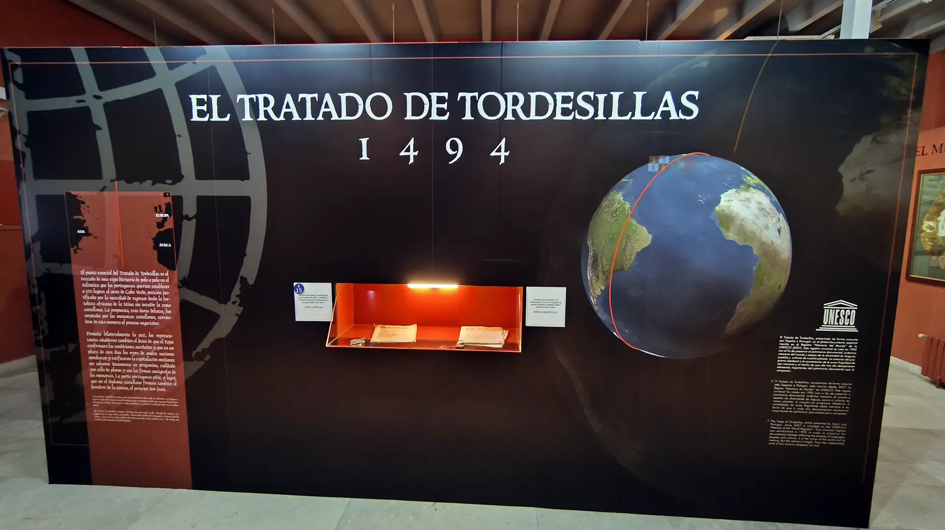 Painel expositivo no Museu do Tratado de Tordesilhas na Espanha com um globo terrestre ilustrando a linha divisória de 1494 entre Portugal e Espanha, documentos históricos em uma vitrine iluminada e o selo da UNESCO.