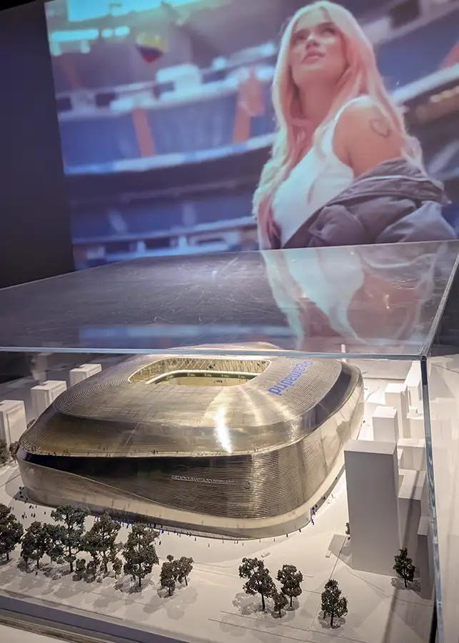 Maquete do Estádio Santiago Bernabéu em exposição numa vitrine com um ecrã gigante ao fundo que mostra uma imagem da cantora Karol G no estádio.