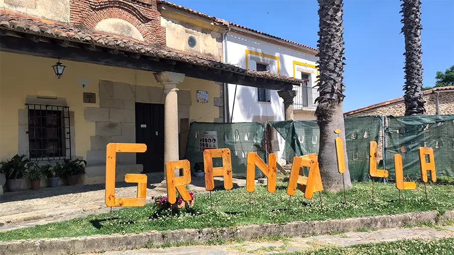 Letras de madeira no gramado formando a palavra Granadilla em frente a um edifício com fachada amarela e varanda.