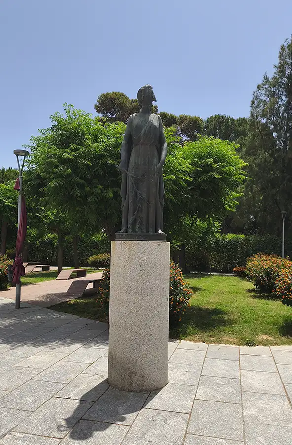 Estátua de bronze em pedestal de granito num jardim público de Mérida, com árvores ao fundo e céu azul