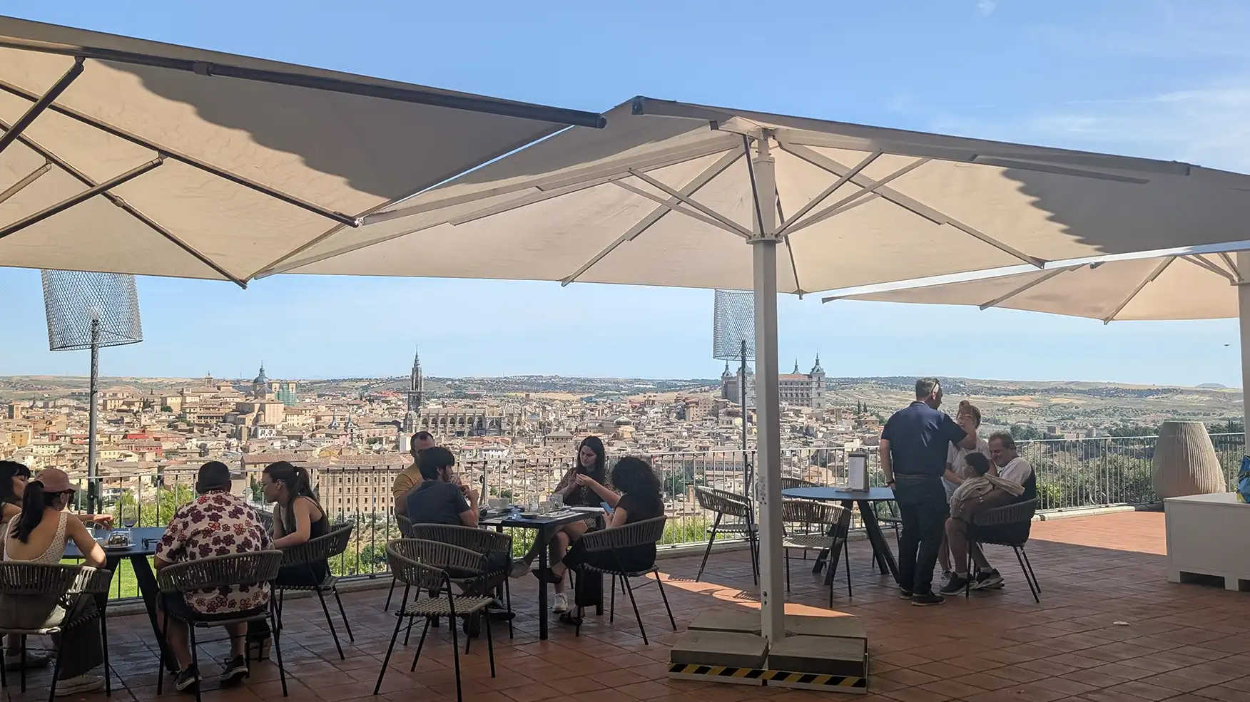 Personas sentadas en mesas de una terraza al aire libre con sombrillas blancas y una vista panorámica de la ciudad histórica de Toledo de fondo.