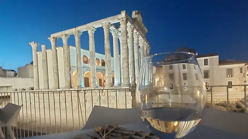 Taça de vinho numa esplanada com vista noturna para o Templo de Diana de Mérida iluminado, no centro histórico da cidade