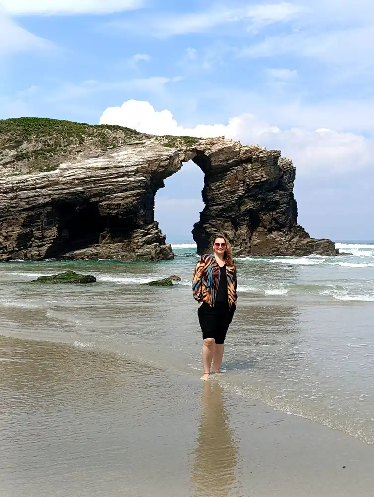 Mulher de pé na beira do mar na praia das Catedrais com grande arco de rocha natural ao fundo
