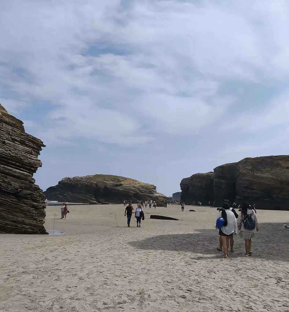 Turistas caminhando pela areia da praia das Catedrais com grandes falésias de rocha em estratos ao fundo