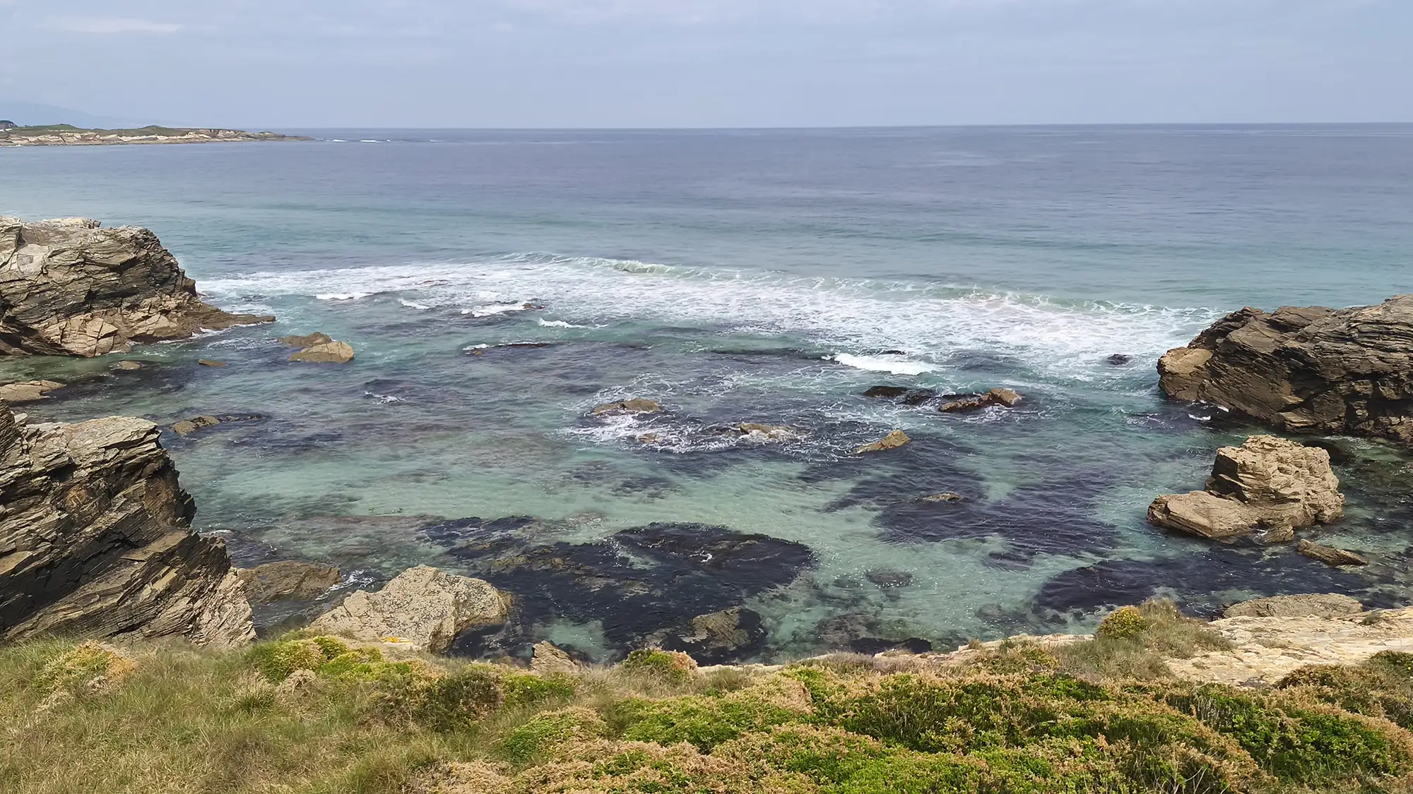Enseada com rochas escuras e água verde-clara na praia das Catedrais com vegetação costeira em primeiro plano