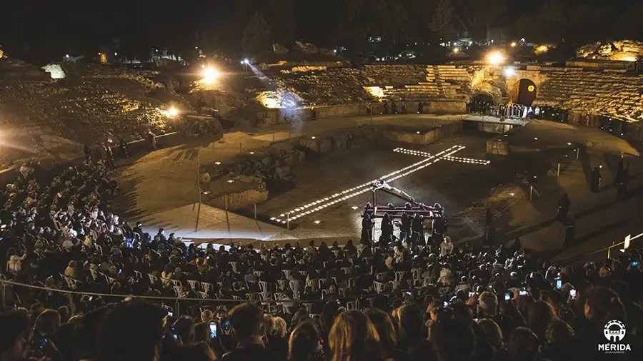 Vía Crucis noturno no Anfiteatro Romano de Mérida durante a Semana Santa, com uma cruz iluminada no centro da arena e o público ao redor