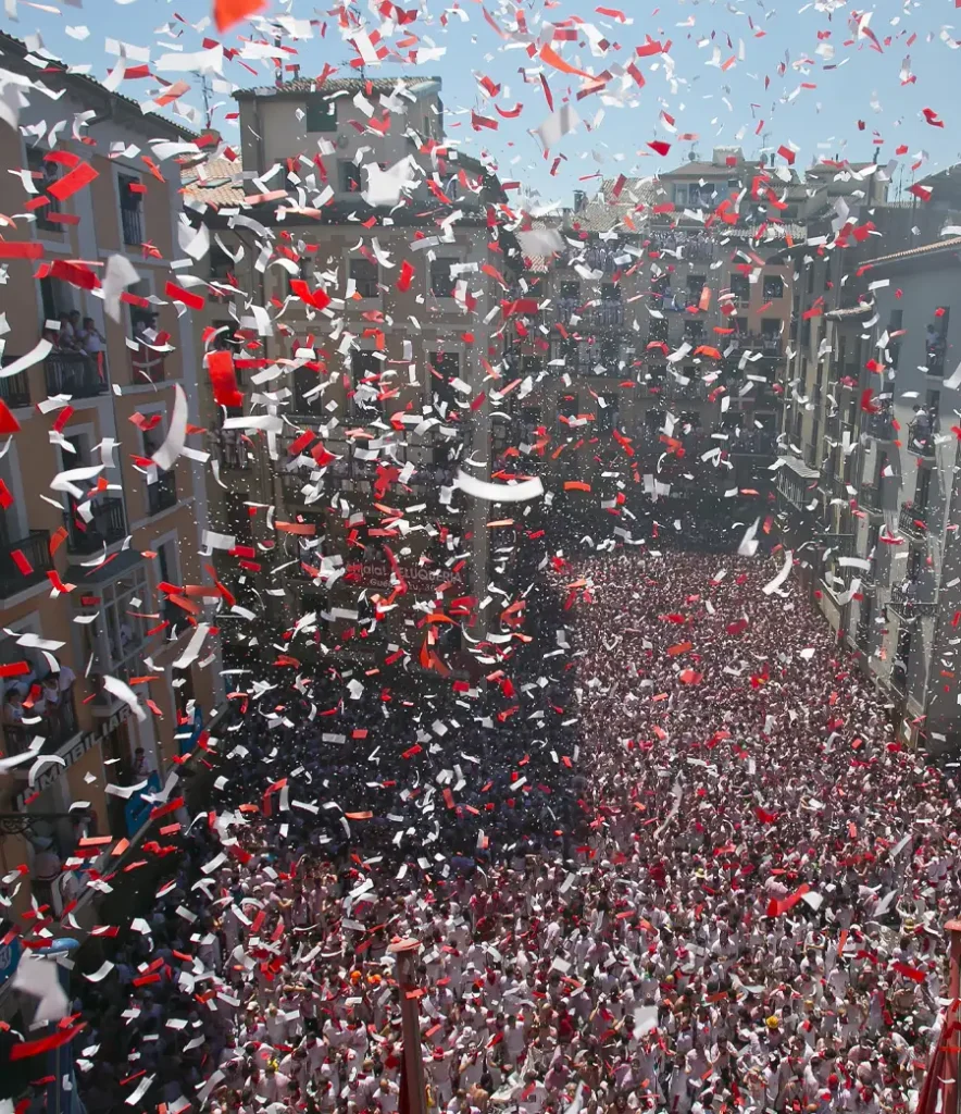 Vista aérea do chupinazo em Pamplona com confete vermelho e branco caindo sobre uma multidão que lota a Plaza Consistorial no início de San Fermín