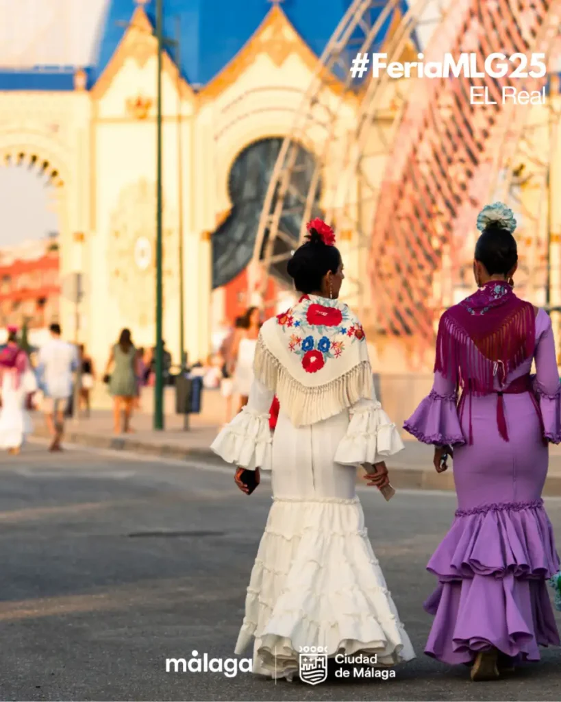 Duas mulheres de vestido de flamenca branco e lilás caminham de costas em direção à entrada iluminada do Real del Cortijo de Torres, o recinto da Feria de Málaga