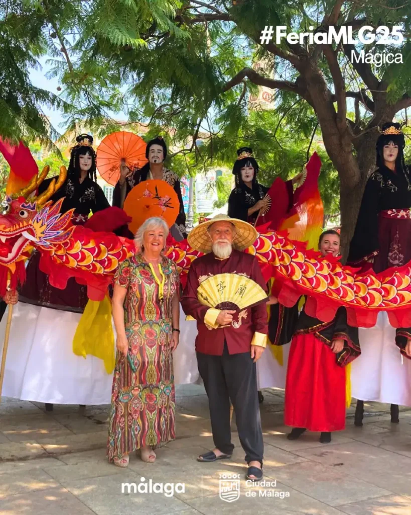 Casal posa com um dragão chinês de tecido e artistas em pernas de pau caracterizados durante a programação infantil da Feria Mágica em Málaga
