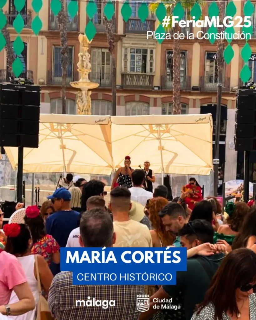 Público lotando a Plaza de la Constitución durante a feira de dia em Málaga, com bandeirolas verdes penduradas e uma cantora se apresentando no palco montado ao lado da fonte