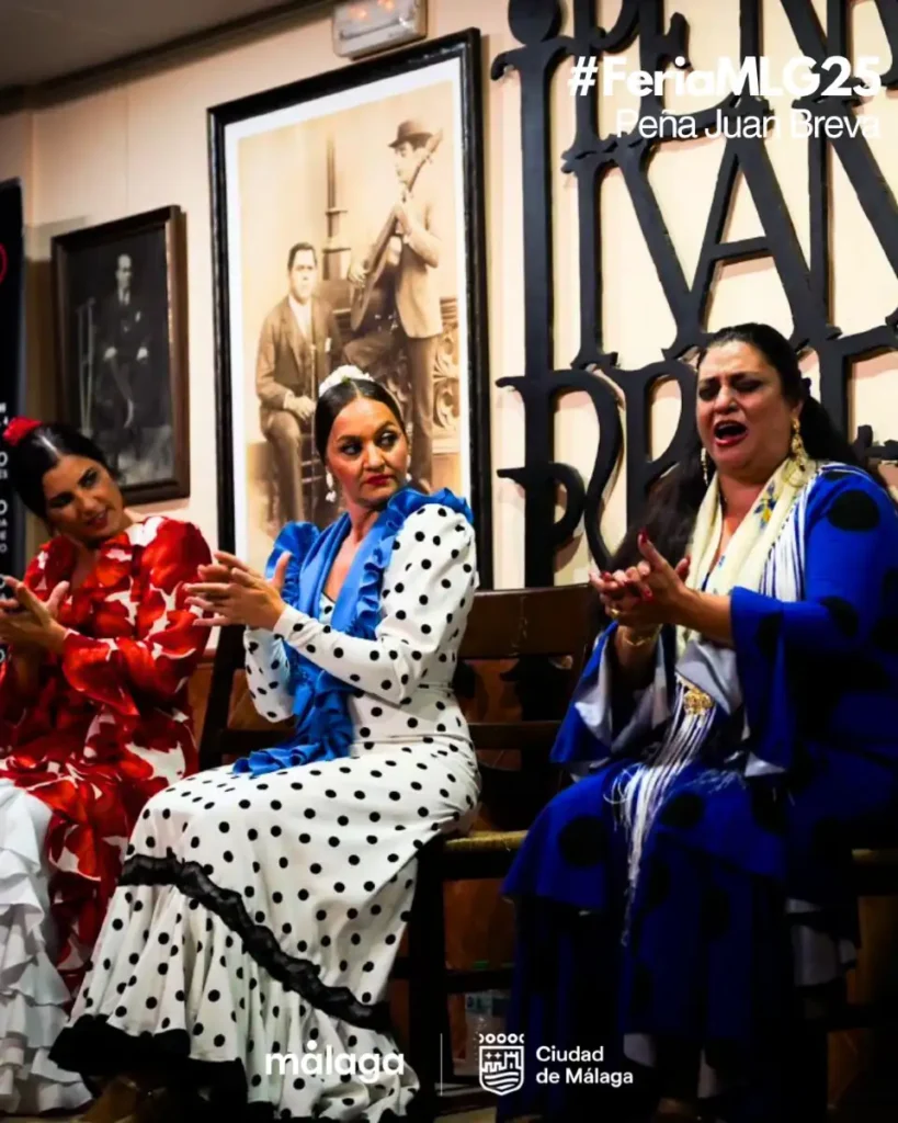 Três mulheres com trajes de flamenca em vermelho, branco de bolinhas e azul cantam e batem palmas sentadas na Peña Juan Breva durante a Feria de Málaga