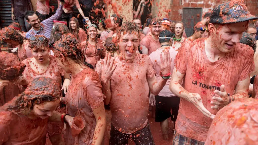 Grupo de participantes com camiseta e corpo cobertos de tomate em uma rua estreita de Buñol durante a Tomatina, com um deles de boca aberta no centro da foto