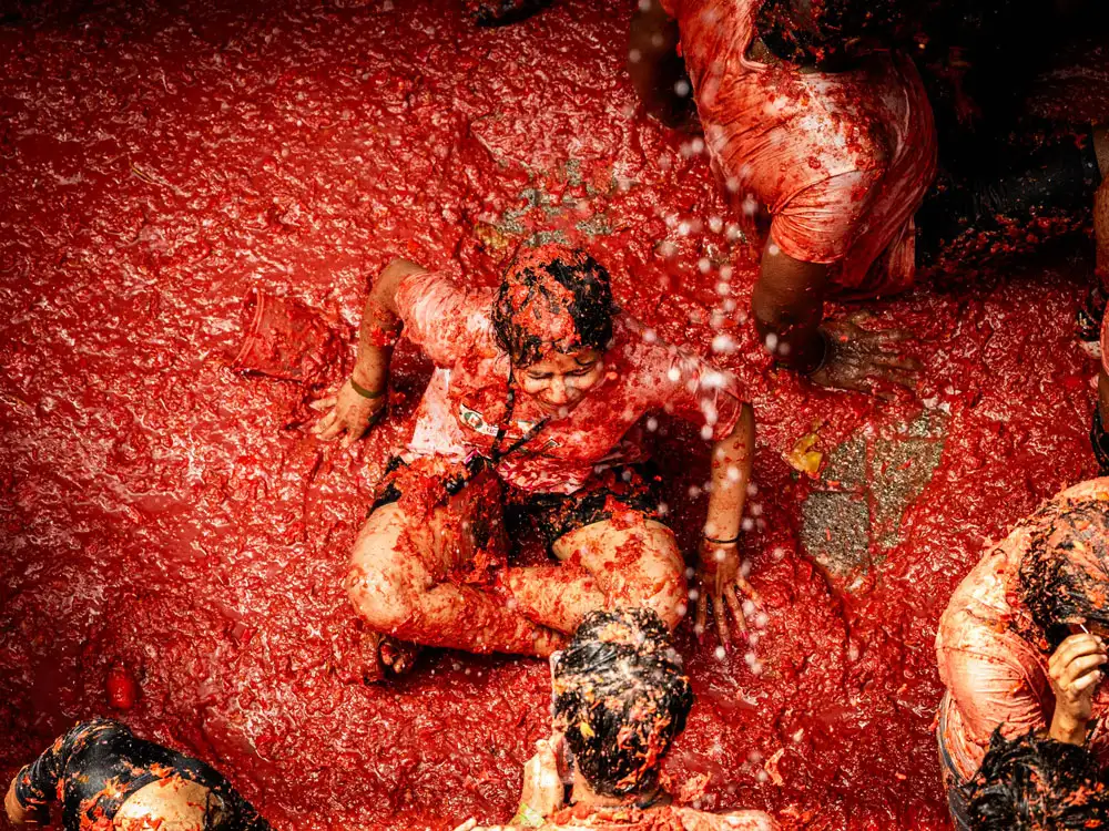Participante sentado no chão coberto por uma camada espessa de polpa de tomate durante a Tomatina, com jatos de água caindo sobre ele