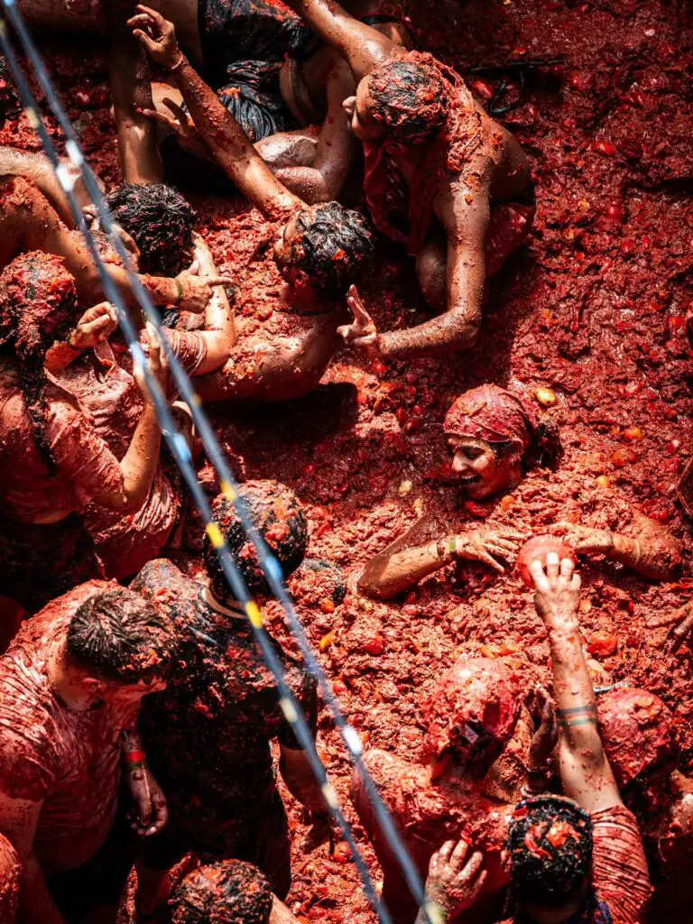Vista de cima da multidão coberta de polpa de tomate durante a Tomatina, com uma participante sorrindo deitada no meio da massa vermelha que cobre a rua