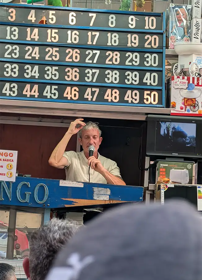 Hombre presentando un bingo en una caseta de las fiestas de Madrid, con el panel de números y micrófono en mano.