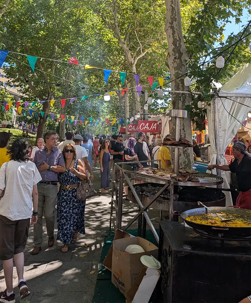 Barraca de comida en las Fiestas de Agosto en Madrid con paella y carne a la parrilla, calle decorada con banderines de colores.