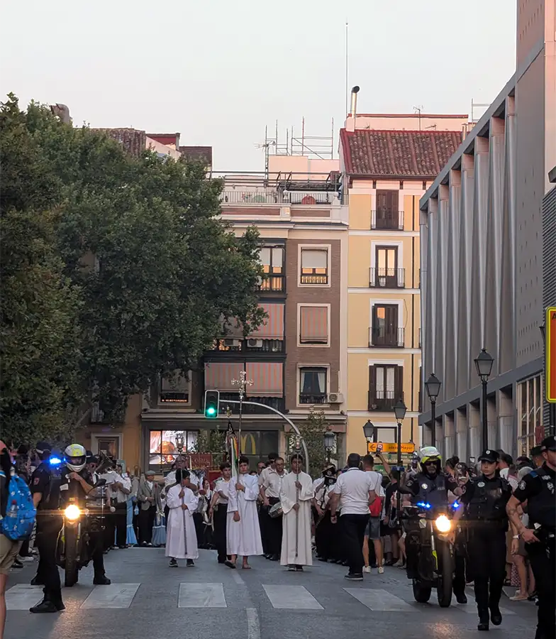Procesión religiosa de las fiestas de agosto en Madrid, participantes vestidos de blanco con una cruz, escoltados por la policía.