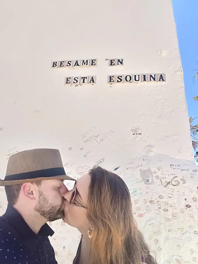 Casal se beijando em esquina de Rota, Espanha, com o azulejo tradicional 'Bésame en esta esquina' na parede branca do centro histórico