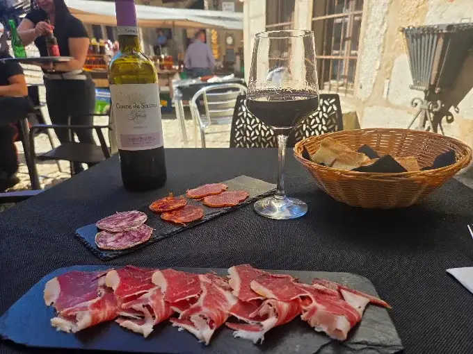 Tábua de embutidos ibéricos e presunto com uma taça de vinho tinto e cesta de pão em uma mesa ao ar livre em La Alberca
