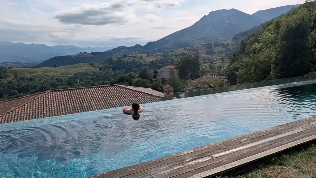 Piscina infinita con vista a un pueblo de montaña y cadena montañosa al fondo, mujer bañándose de espaldas en primer plano