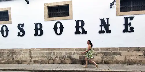 Mulher caminha em frente à fachada histórica da bodega Osborne com o nome em letras grandes.