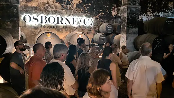 Um grupo de turistas visita uma adega histórica da Osborne, com barris empilhados e um letreiro luminoso 'OSBORNE Y Ca. FOUNDED IN 1772'.