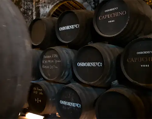 Barris de xerez empilhados na bodega exibindo o logotipo da Osborne e a marcação Capuchino VORS em letras brancas.