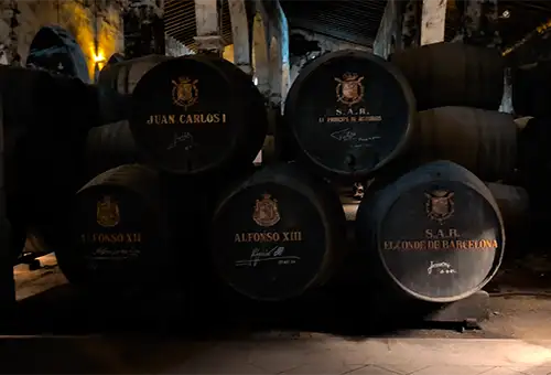 Pilha de barris escuros exibindo brasões e nomes de membros da realeza espanhola como Juan Carlos I e Alfonso XIII.