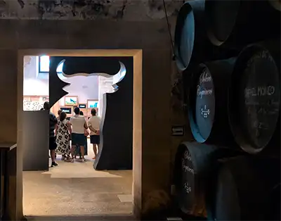 Turistas passam da adega escura para uma galeria iluminada através de um pórtico com o recorte da cabeça de um touro.