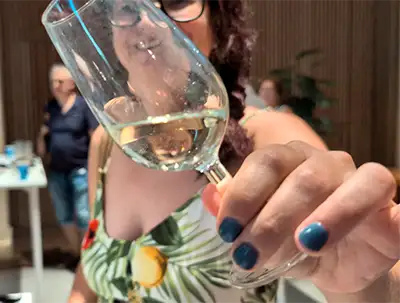 Mulher sorridente com óculos e blusa floral segura uma taça de vinho branco na direção da lente durante a degustação.