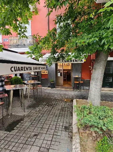 Fachada do Cuarenta Bar de Vinos, com mesas altas do lado de fora sob guarda-sol com o nome do bar, entre árvores e calçada de pedra.