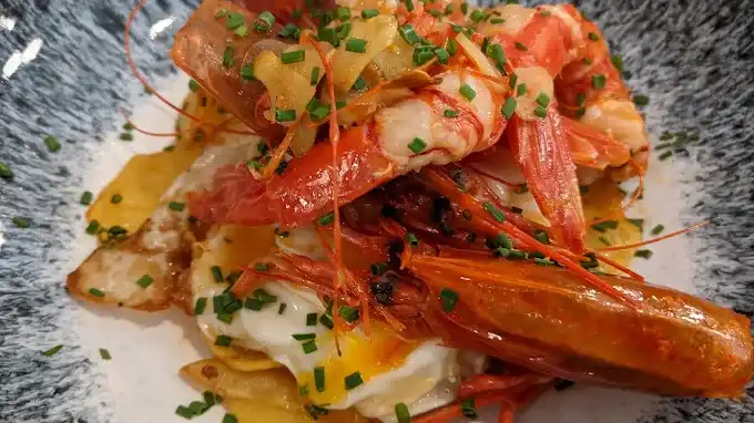 Prato com camarões vermelhos grelhados sobre batatas fritas e ovo com a gema mole, finalizado com cebolinha picada.