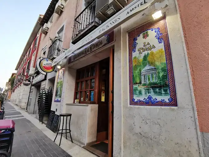 Fachada da taberna La Tinaja em uma rua de pedestres de Aranjuez, com painel de azulejos com um pavilhão de jardim ao lado da porta de madeira e toldo com o nome do bar.
