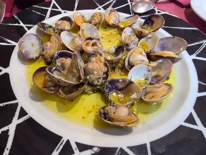 Prato com amêijoas abertas em molho de azeite com alho e salsa, sobre mesa de tampo preto com desenho geométrico branco.