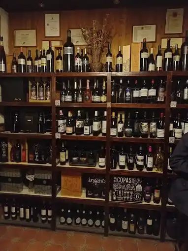 Estante de madeira cheia de garrafas de vinho e licores, com diplomas emoldurados na parede ao fundo.