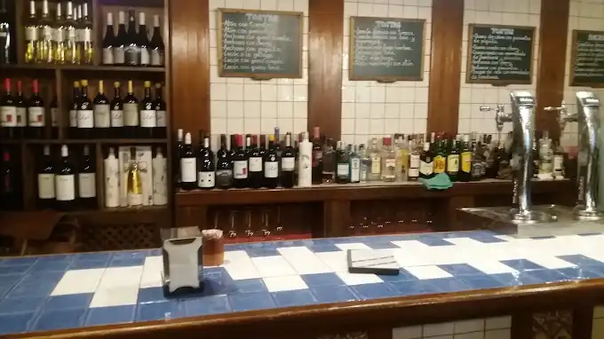 Balcão com tampo de azulejos azuis e brancos, prateleiras de madeira com garrafas de vinho e licores, chopeiras de metal à direita e lousas com o cardápio de tostas na parede.