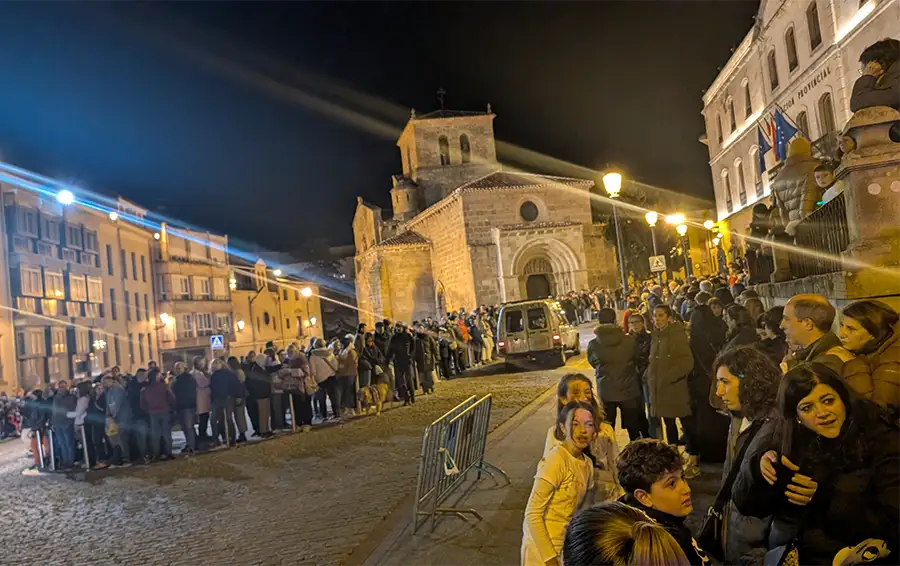 Multidão de pessoas aglomerada atrás de grades de contenção aguardando o desfile noturno do Festival de las Ánimas em Soria com a Igreja de San Juan de Rabanera iluminada ao fundo.