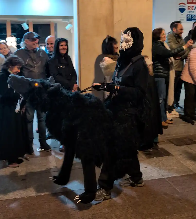 essoa com máscara branca e capuz preto a conduzir uma criatura peluda escura pelas ruas de Soria.