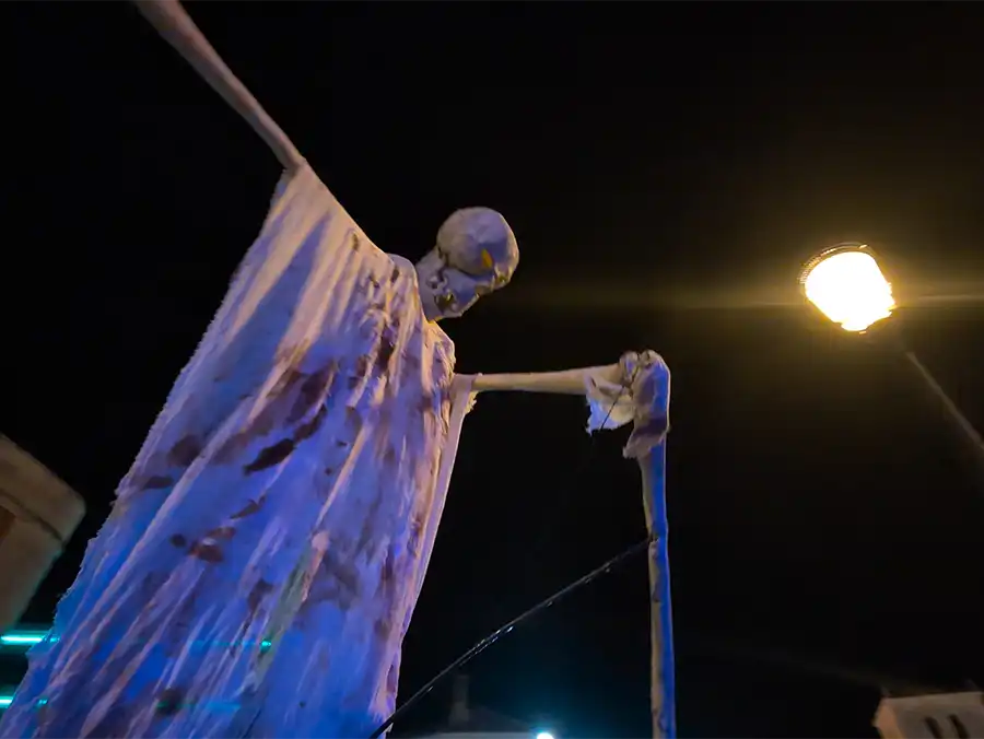 Figura spectral de caveira de grandes dimensões com túnicas brancas e esfarrapadas em desfile noturno, vista de ângulo baixo sob a luz de um poste de rua.
